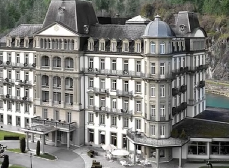 Historic façade of Grand Hotel Beau Rivage in Interlaken, Switzerland, situated by the calm Aare River with mountain peaks in the background under a bright sky.